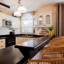 Open‑plan dining space in the Portofino two‑bedroom apartment, showing a table with chairs and a fully equipped kitchen behind, including refrigerator/freezer, dishwasher, oven, cooktop, microwave, air‑fryer, electric kettle and Nespresso coffee machine.
