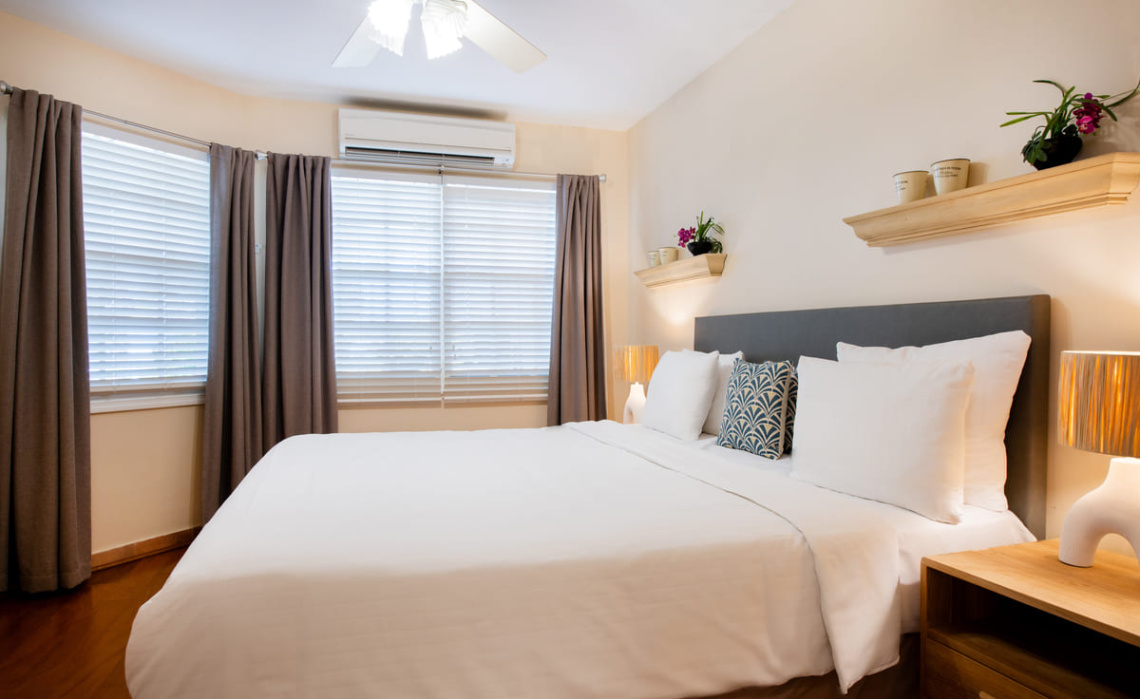 Primary bedroom of the Portofino apartment featuring a king‑size bed, walk‑in closet and en‑suite bathroom, with blinds, curtains, air‑conditioning and ceiling fan.