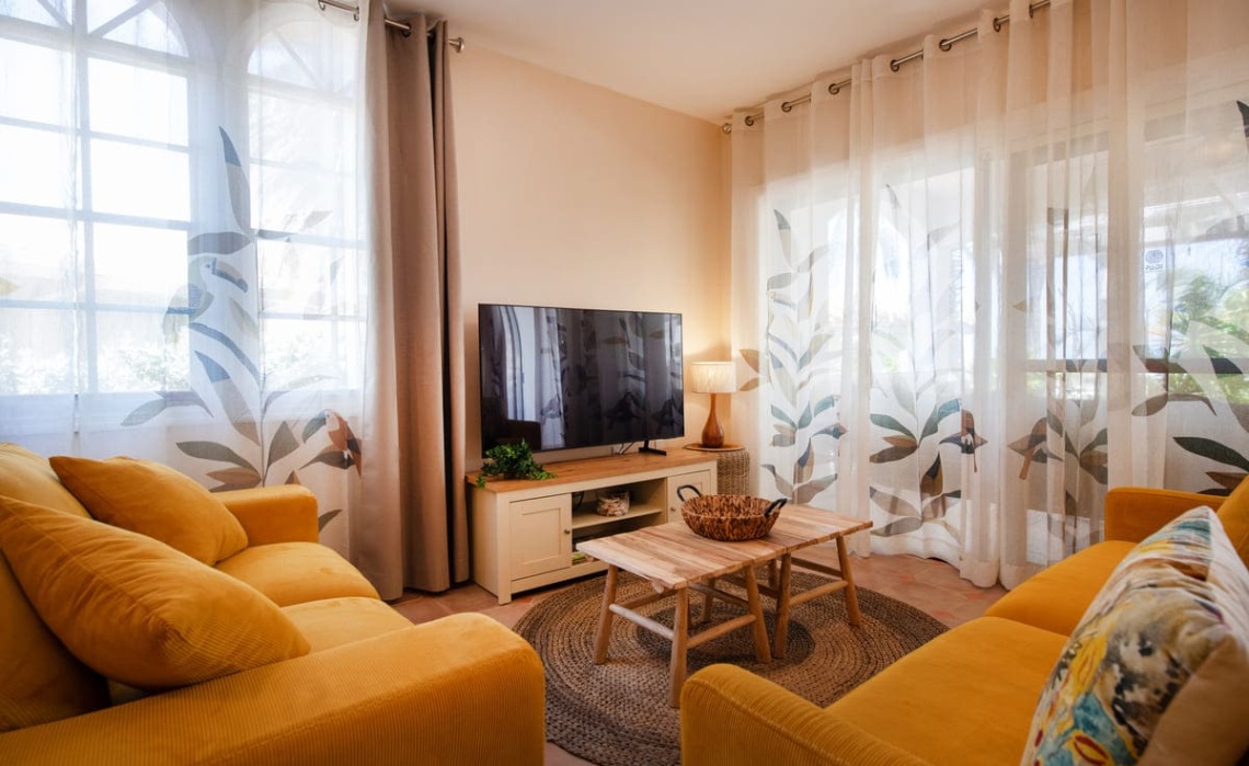 Living room of the Portofino two‑bedroom apartment showing two sofas in vivid colours, a wall‑mounted Smart TV, and assorted decorative accents such as cushions, a coffee table and decorations.