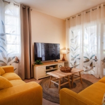 Living room of the Portofino two‑bedroom apartment showing two sofas in vivid colours, a wall‑mounted Smart TV, and assorted decorative accents such as cushions, a coffee table and decorations.