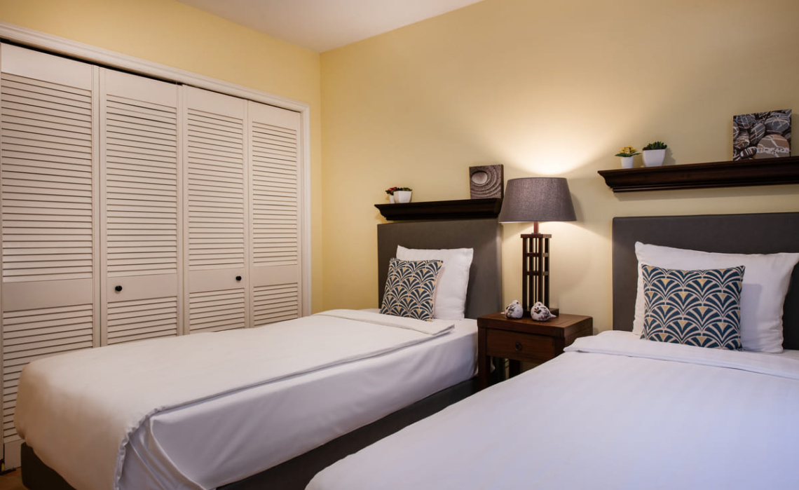 Second bedroom of the Portofino apartment featuring two single beds that can be joined, built‑in closet and additional storage space, with air‑conditioning, ceiling fan, blinds and curtains.