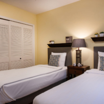 Second bedroom of the Portofino apartment featuring two single beds that can be joined, built‑in closet and additional storage space, with air‑conditioning, ceiling fan, blinds and curtains.